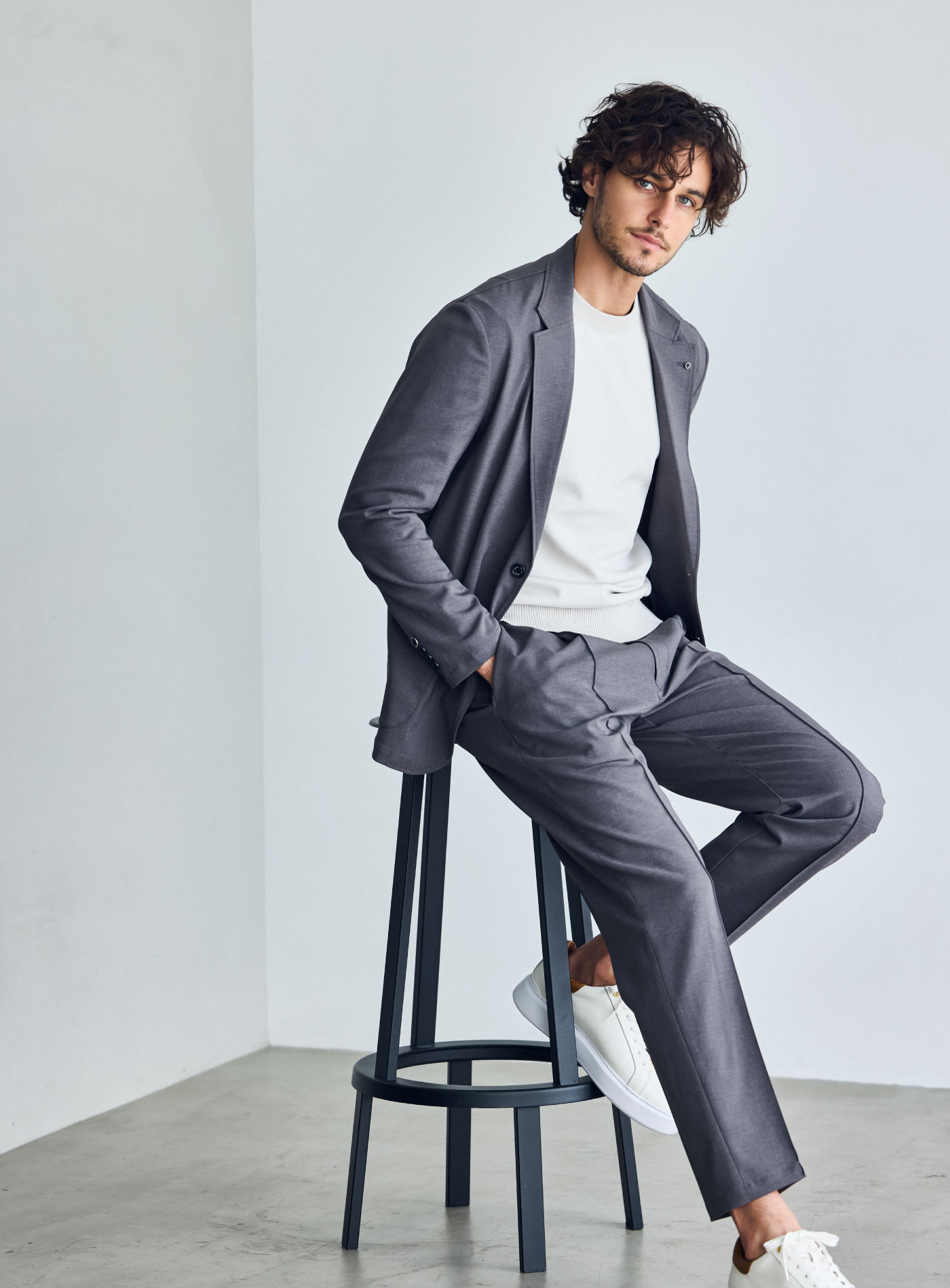 ライトブラウンのスーツ・アイボリーのニット・ホワイトのスニーカーを着用した、黒の椅子に寄りかかっている男性