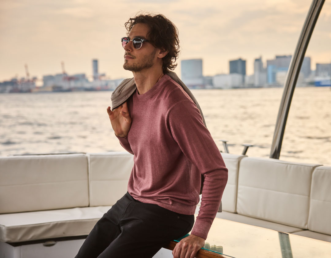 ライトピンクのトップス・アイボリーのジャケット・ブラックのデニムパンツを着用した男性がジャケットを方に乗せて船の椅子に座っている様子