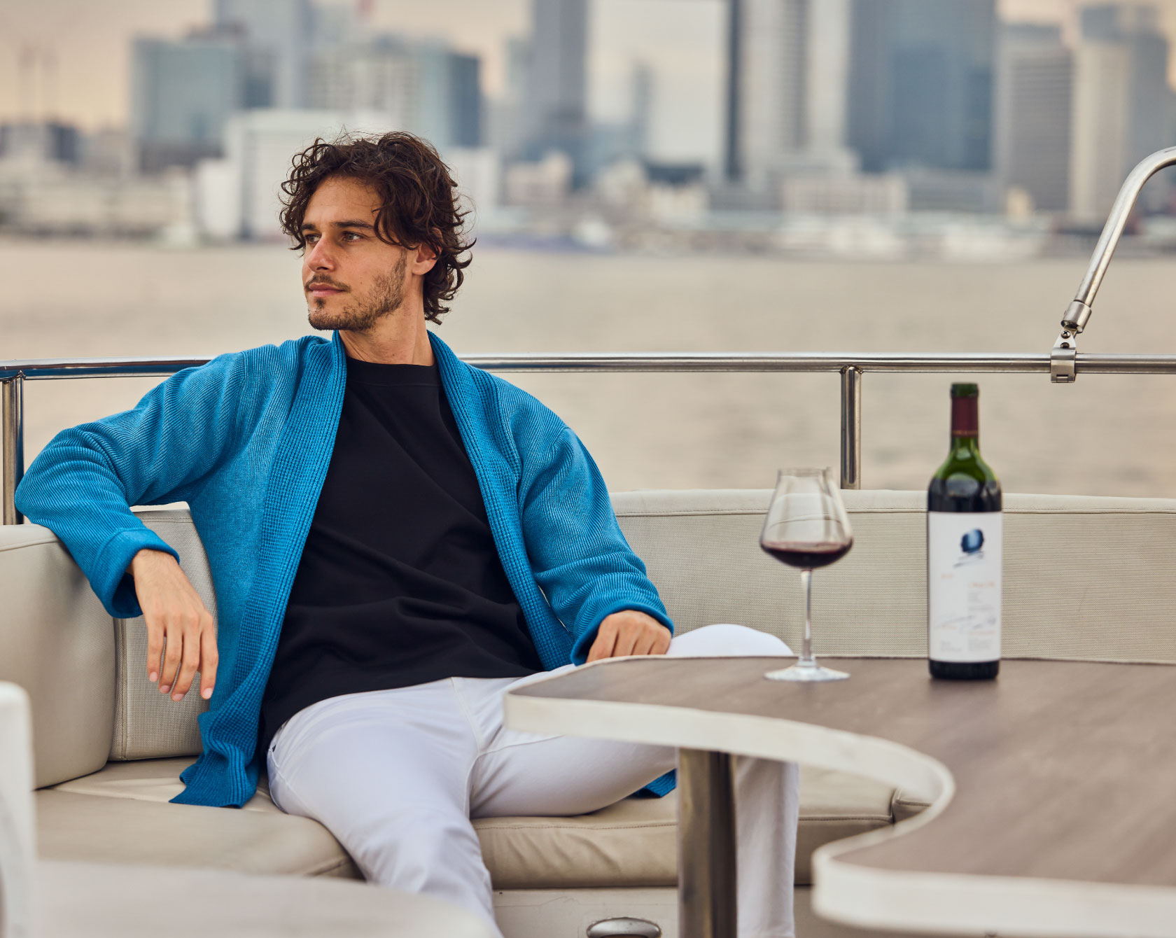 ブルーのカーディガン・ブラックのトップス・ホワイトのデニムパンツを着用した男性が船の白いソファに座って遠くを眺めている様子