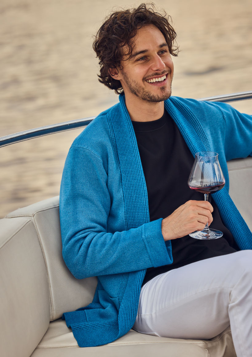 ブルーのカーディガン・ブラックのトップス・ホワイトのデニムパンツを着用した男性がワインを持って船の白いソファに座って笑っている様子