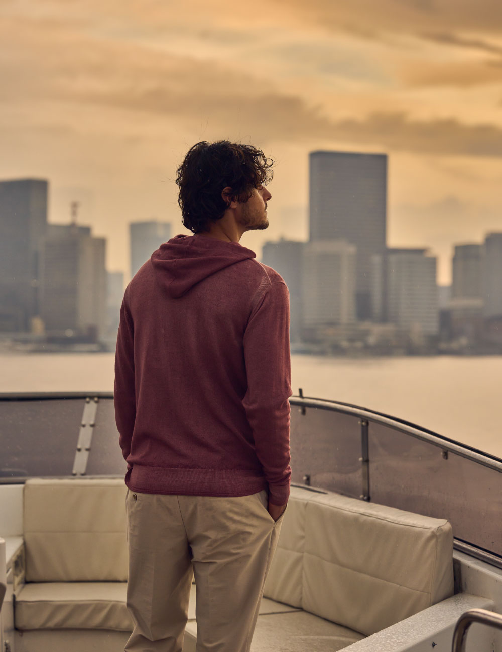 ライトピンクのパーカー・ライトベージュのパンツを着用した男性が船の上で遠くを眺めている様子