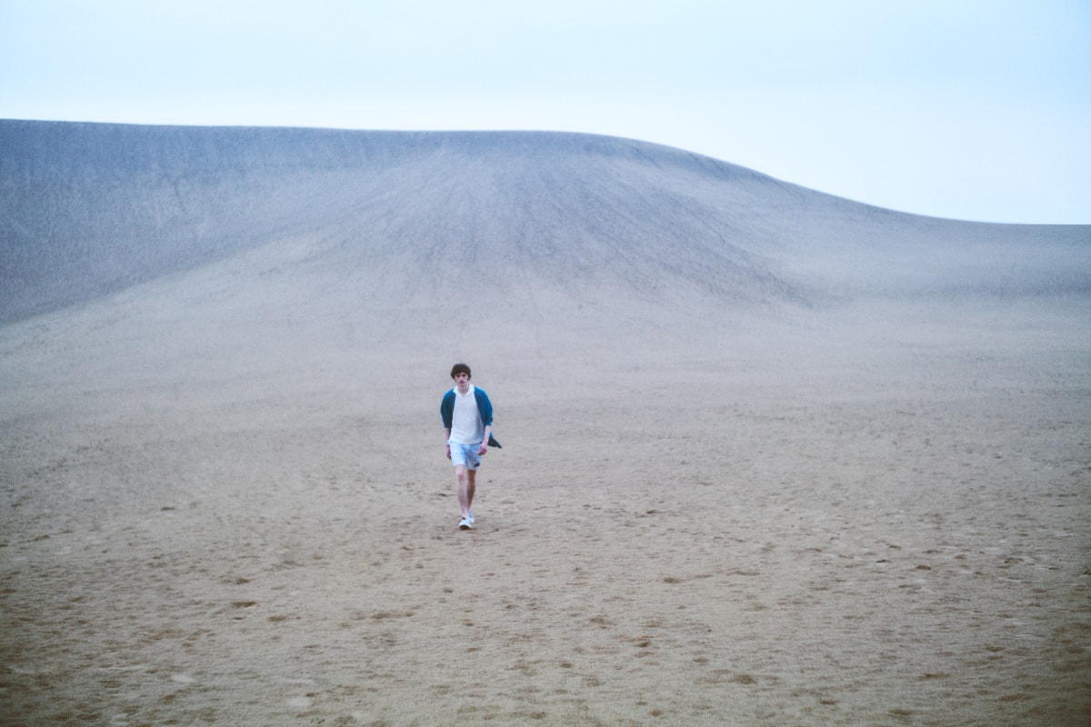 青いカーディガンにショートパンツを着た男性が砂漠を歩いている画像